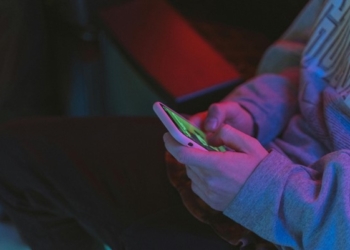 Uma Pessoa Branca De Casaco Mexendo No Celular Com As Duas Mãos. A Foto Só Foca Nas Suas Mãos E Braço, Não Exibindo Claramente O Restante Do Seu Corpo.